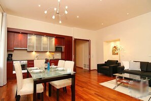 A modern living room with a glass dining table, dark sofas, and a kitchen in the background.