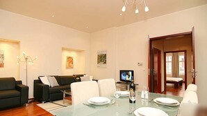 Un salon moderne avec une table à manger en verre pour quatre personnes, un canapé noir, un téléviseur et des œuvres d’art accrochées aux murs.