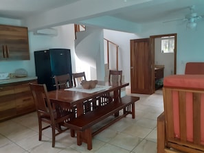 Un comedor con una mesa y sillas de madera, un banco, una cocina con refrigerador y una puerta que da a otra habitación.