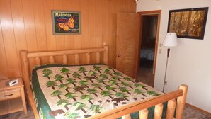 A bedroom with a wooden bed frame, a quilt with a nature pattern, a Mariposa apples poster, a lamp, and a door leading to another room. - End of the road .... (Mariposa)