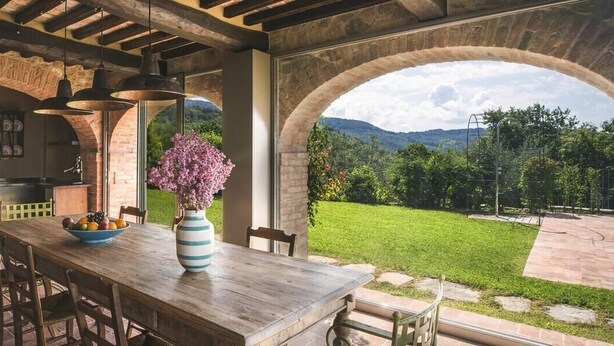 An outdoor kitchen with a brick fireplace, a dining table with chairs, and a view of a green landscape through an archway. - Villa Mandorla - Tuscan farmhouse with private pool (Anghiari)