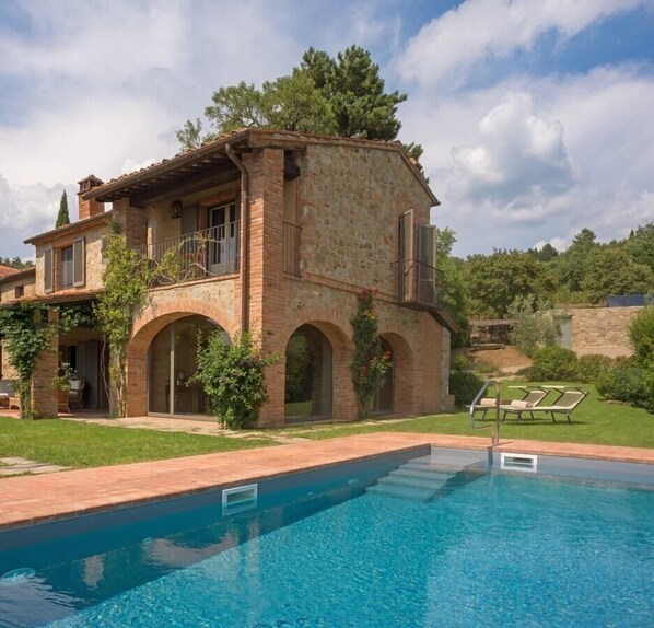 A stone villa with a swimming pool, arched windows, and a garden with lounge chairs.