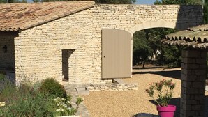 A stone building with a tiled roof, a wooden door, and a small archway. There are two wooden lounge chairs and a potted plant. - Gordes/Goult - Stunning Mas Provençal On A Hill In Luderon (Goult)
