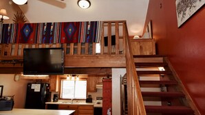 Une chambre de style « loft », avec un escalier en bois, un téléviseur à écran plat fixé au mur et un tapis coloré sur le sol.