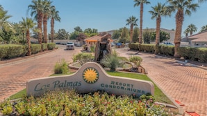 A sign for Las Palmas Resort Condominiums with a sun emblem, surrounded by palm trees and a paved driveway.