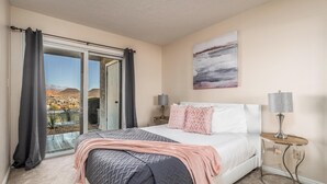 A bedroom with a bed, a nightstand, a lamp, a painting, and a view of a mountainous landscape through a sliding door.
