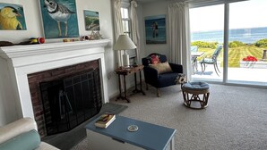 A living room with a fireplace, a blue armchair, a round ottoman, and a view of the sea through large windows.