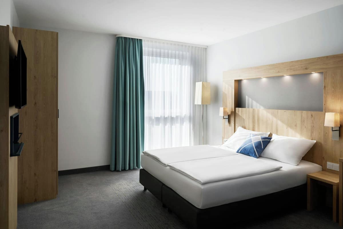 Ein Hotelzimmer mit Bett, Nachttisch, Lampe, Holz-Kopfteil, Fenster mit Vorhängen und Kleiderschrank.