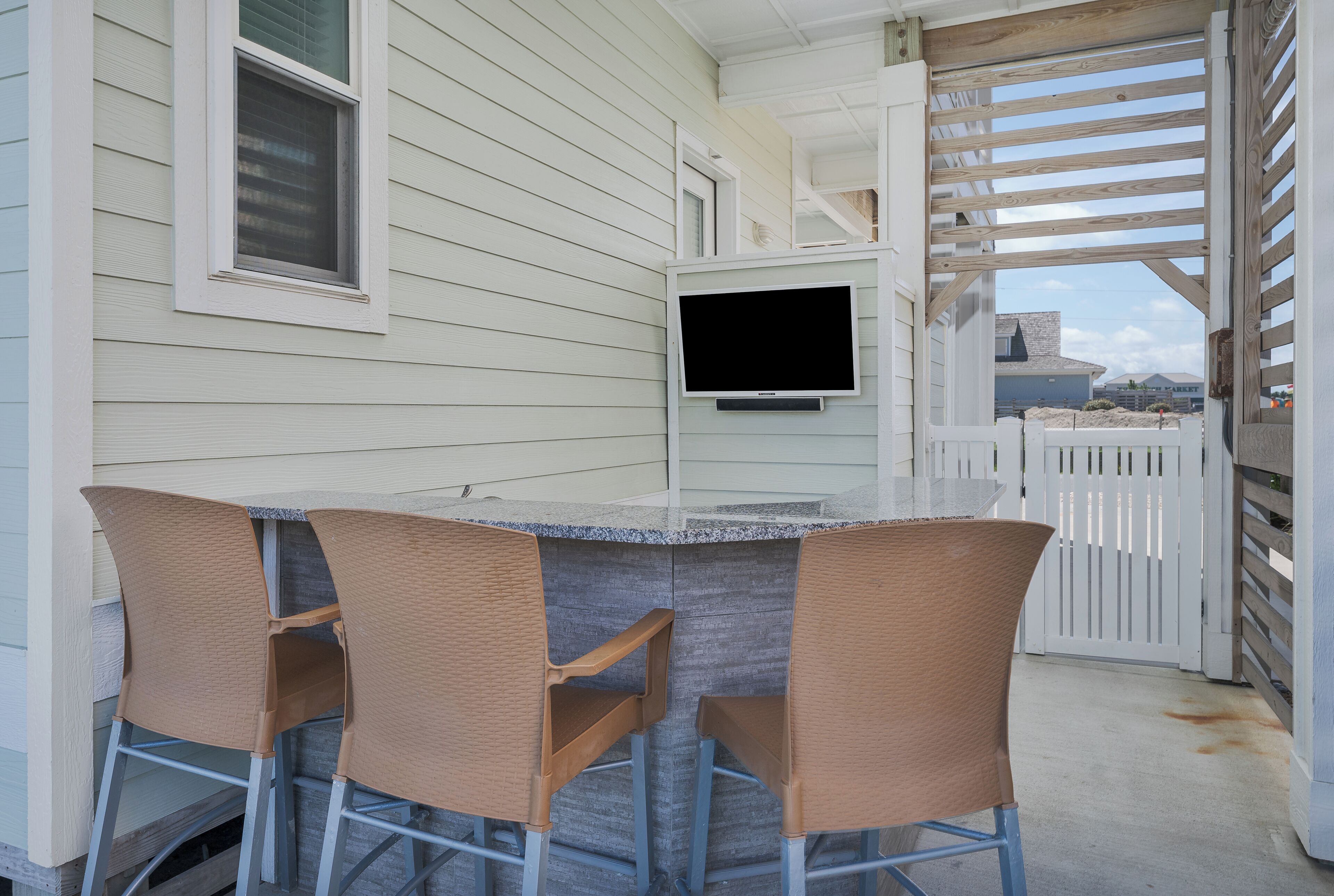 An outdoor patio with a table and chairs, a flat-screen TV mounted on the wall, and a white fence in the background.