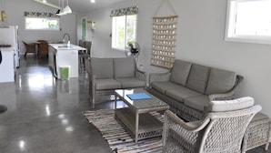 Una sala moderna con un sofá gris, sillones de mimbre, una mesa de centro de cristal y una cocina al fondo.