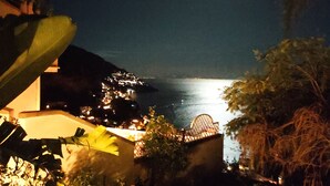 Une scène côtière nocturne, avec une vue sur la mer éclairée par la lune, des bâtiments illuminés et une végétation luxuriante.