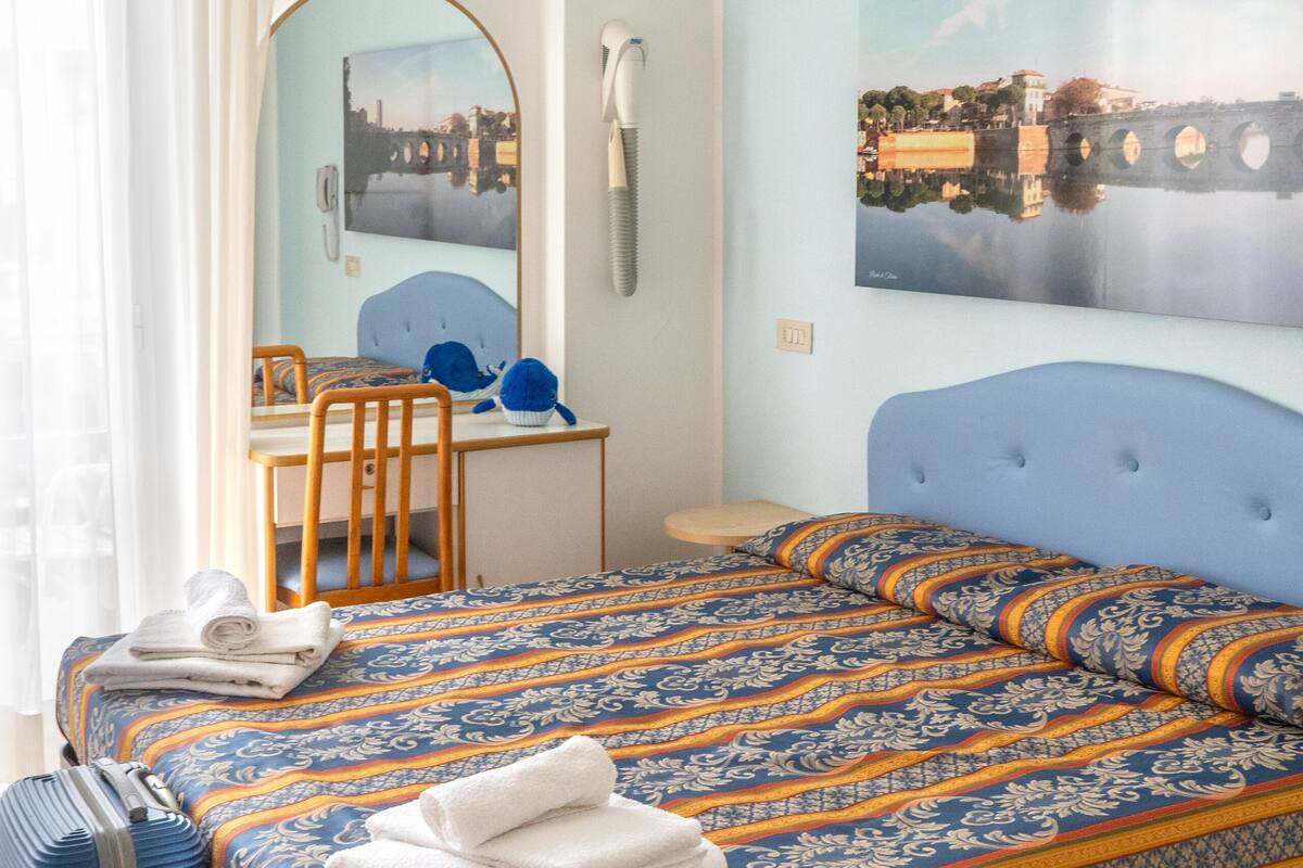 A hotel room with a bed, a desk, a chair, a suitcase, and a painting on the wall.