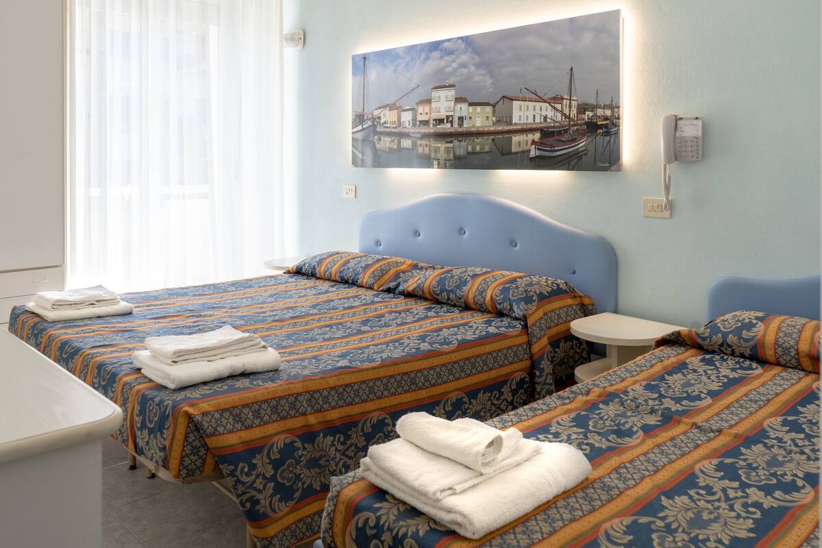 A hotel room with two beds, a small table, and a painting of a harbor scene.