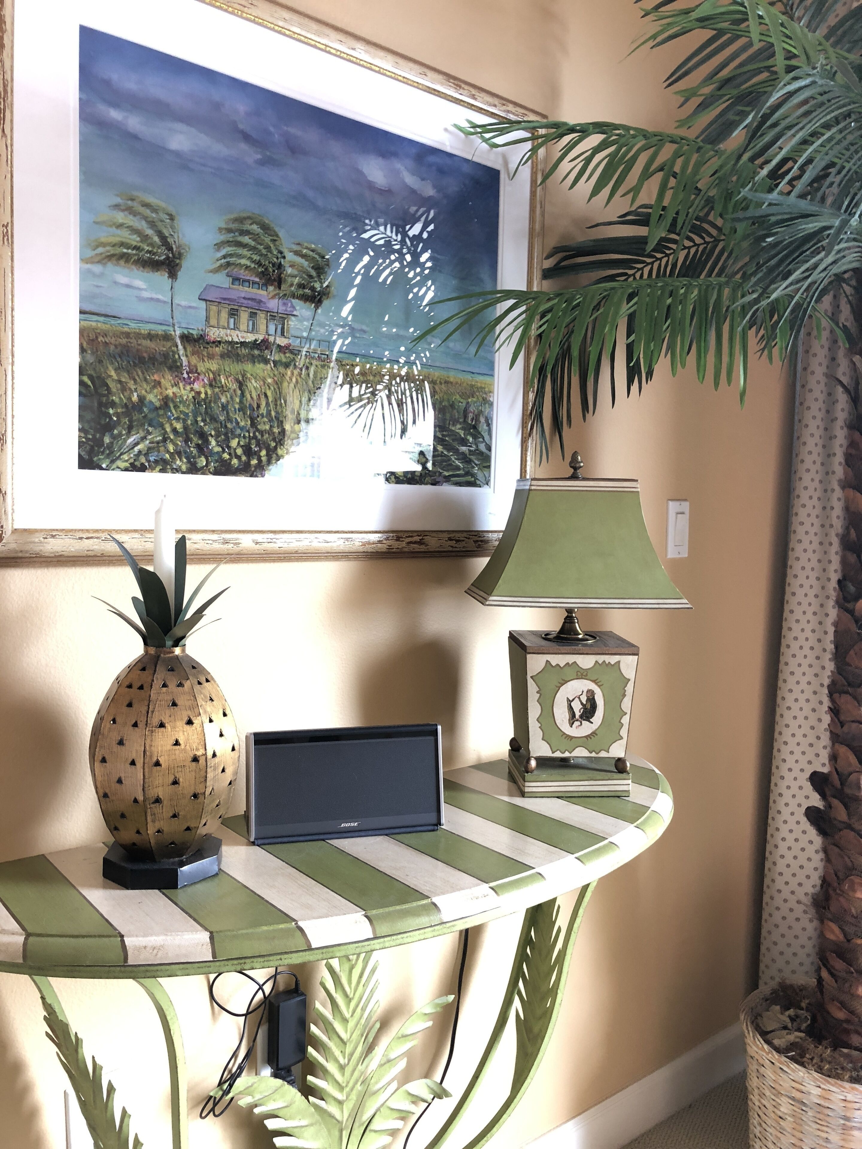 A room with a framed painting of a tropical scene, a pineapple-shaped vase, a lamp, a small table with a laptop, and a potted plant.