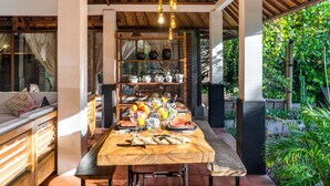 Un comedor con una mesa de madera dispuesta para una comida, rodeado de estantes con utensilios de cocina, y vistas a un follaje exuberante.