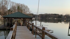 A wooden pier leading to a covered dock area with a green roof, extending over calm water with boats and trees in the background.
