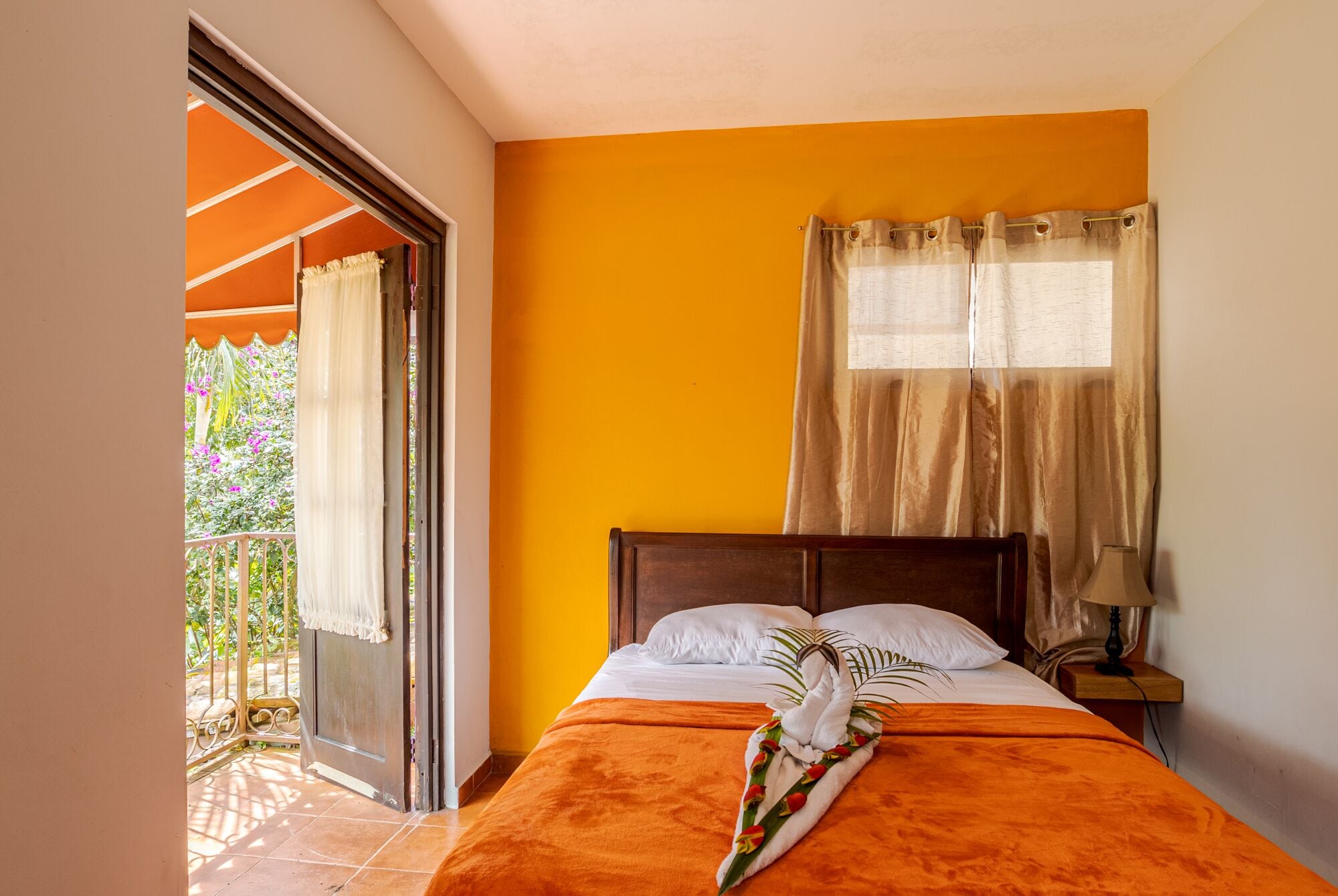 Habitación de hotel con cama, ventana con cortinas, balcón con vista a follaje y puerta que da al exterior.