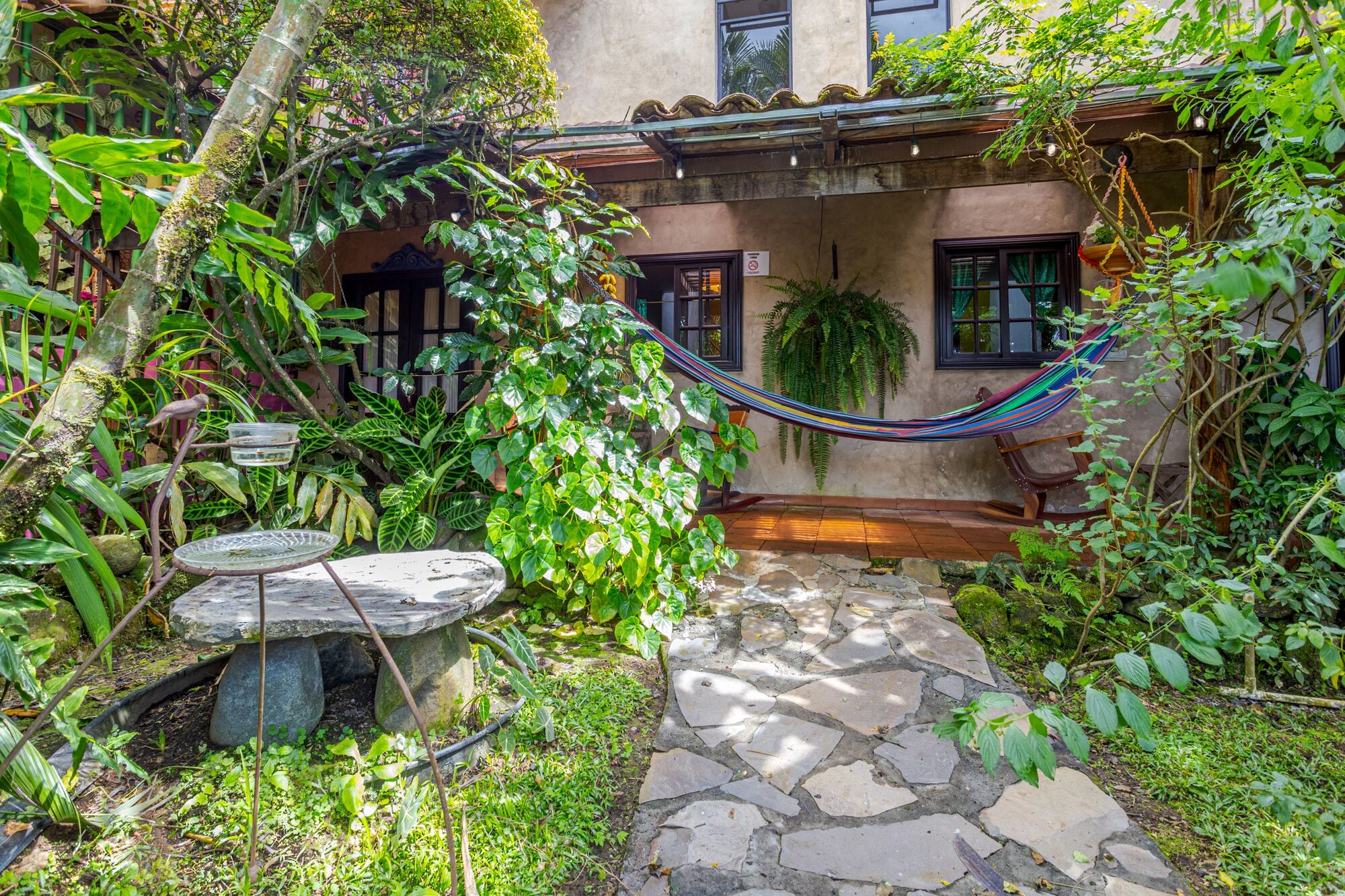 A stone pathway leading to a rustic building with a hammock and a stone bench.