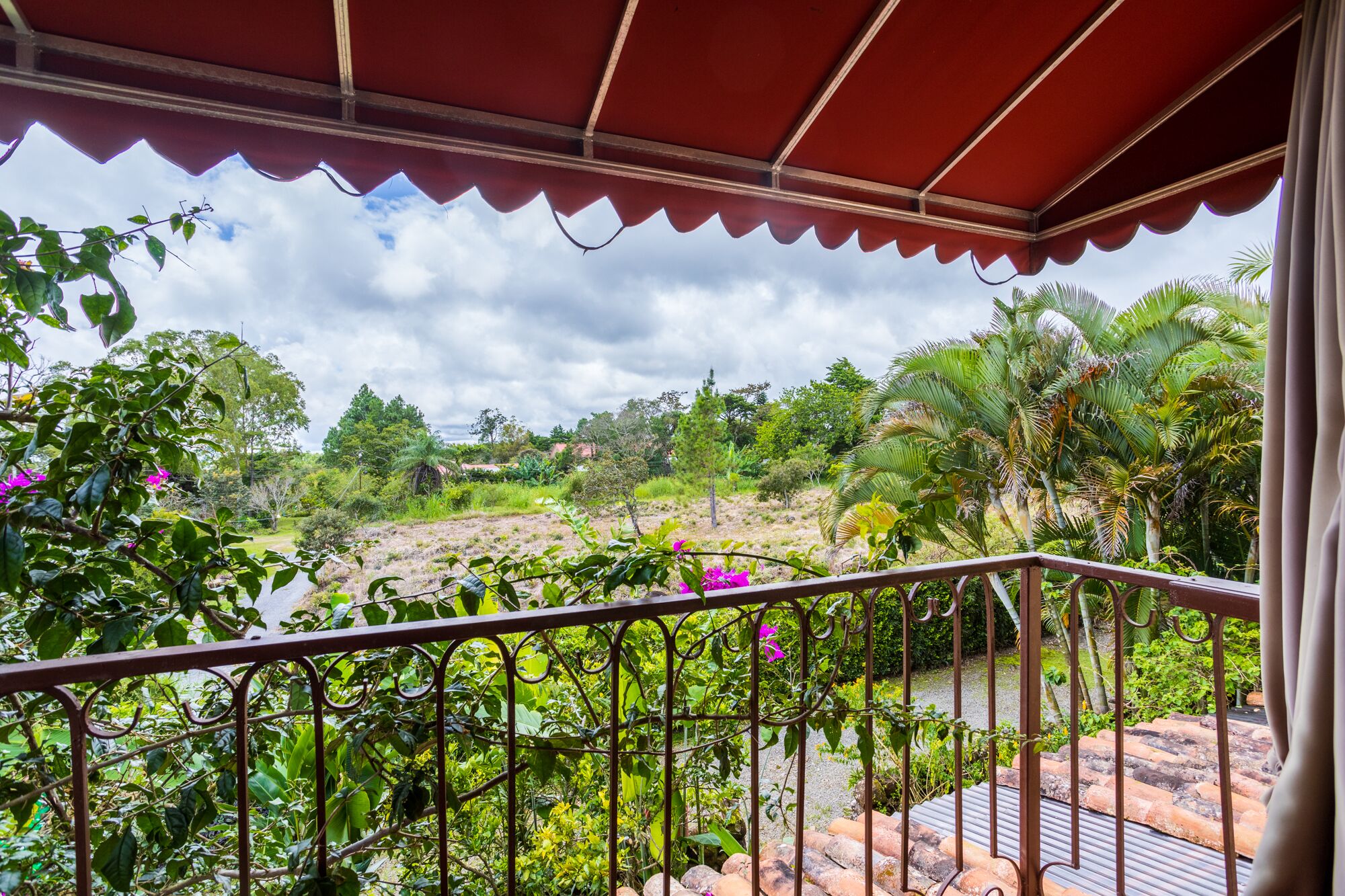 Un balcón con toldo rojo que da a un jardín exuberante con diversas plantas y vistas a un paisaje montañoso.