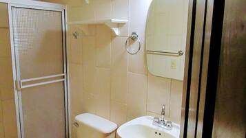Un baño pequeño con inodoro, lavamanos y ducha. Las paredes están cubiertas de azulejos blancos y hay una puerta blanca.