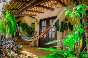 A hammock hanging between wooden beams, surrounded by lush green plants and a tiled floor.