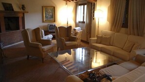 Una sala de estar con una mesa de centro de vidrio, sofás y sillones color beige. Hay una chimenea, un televisor y un cuadro en la pared.