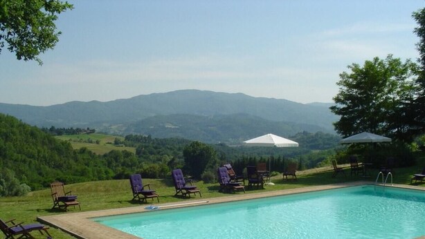 Una piscina rectangular con cubierta de madera, rodeada de sillas de jardín y sombrillas, con colinas y montañas al fondo.