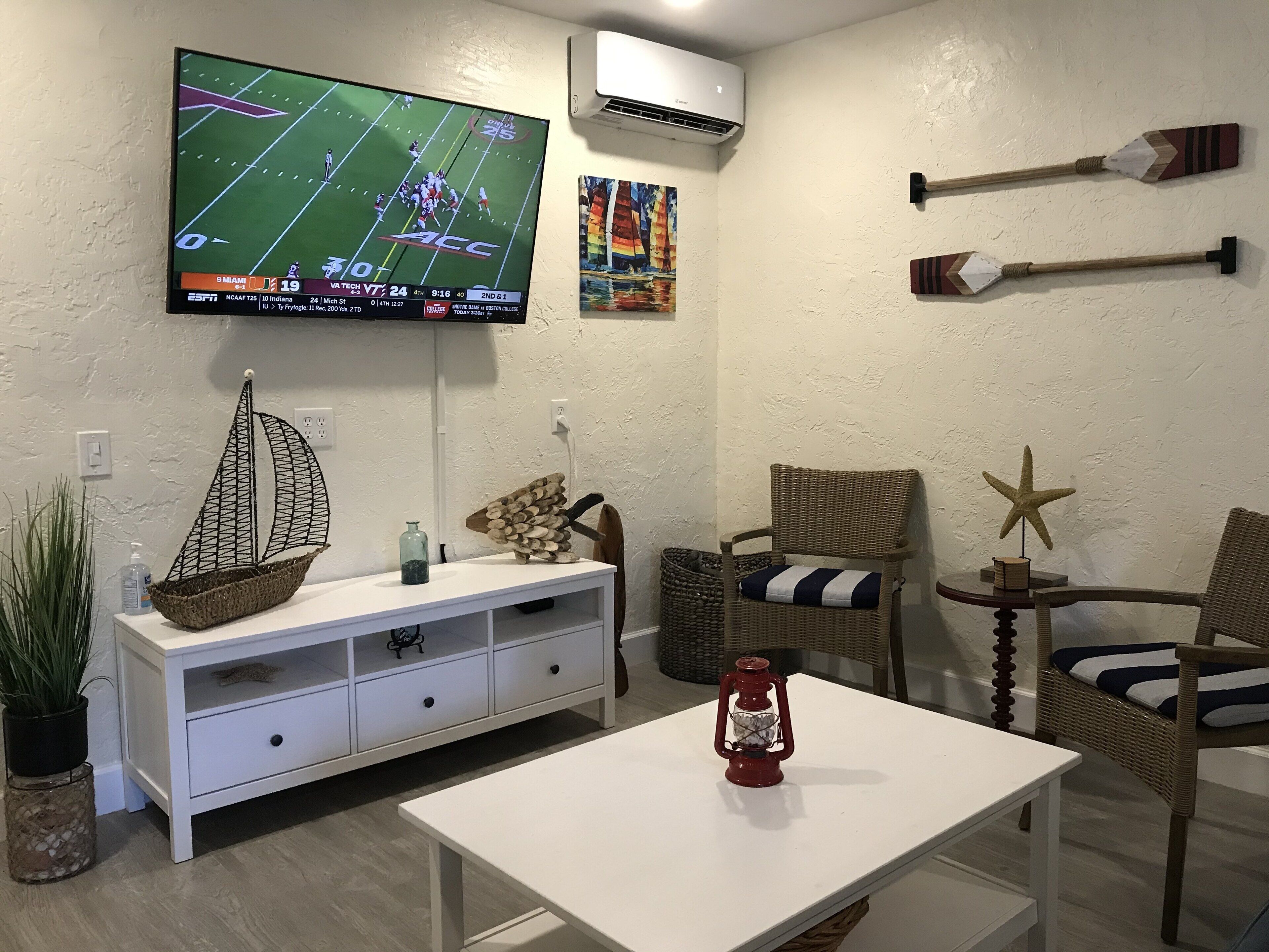 Ein Wohnzimmer mit einem an der Wand befestigten Flachbildfernseher, einem weißen TV-Schrank mit Schubladen, einem Korbstuhl, einem kleinen Tisch mit einer roten Laterne und Wanddekorationen.
