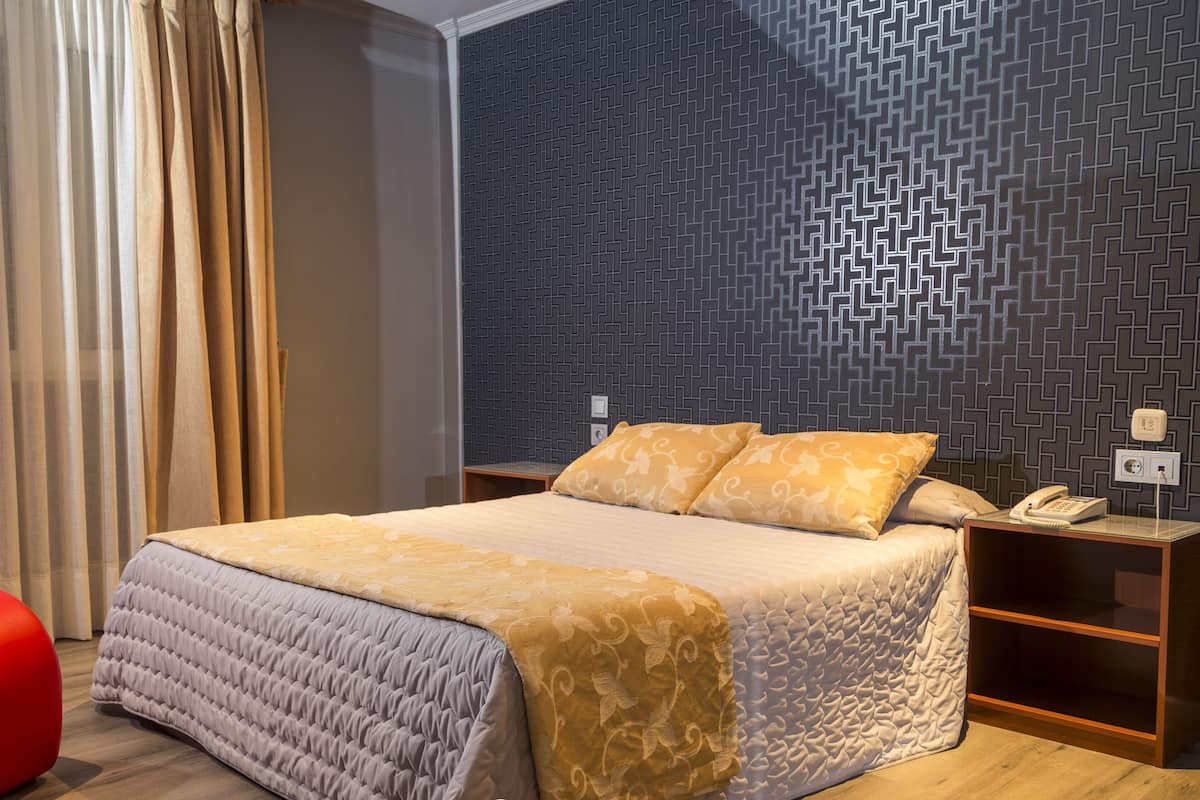 A neatly made bed with a patterned bedspread and two pillows, a bedside table with a telephone, and a wall with a geometric pattern.