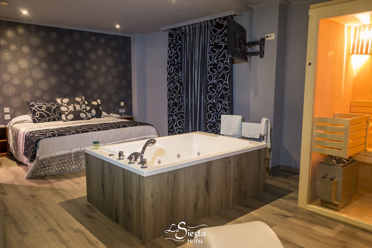 A modern hotel room with a large bathtub, a bed with patterned bedding, and a wooden floor.