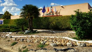 Un edificio con varias banderas, un jardín cuidado con rocas y plantas, y un cielo azul despejado.