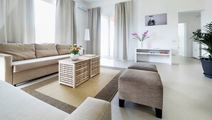 Un salon moderne avec un canapé, un pouf et une table basse. On y trouve une plante en pot et une étagère murale sur laquelle est posée une image.