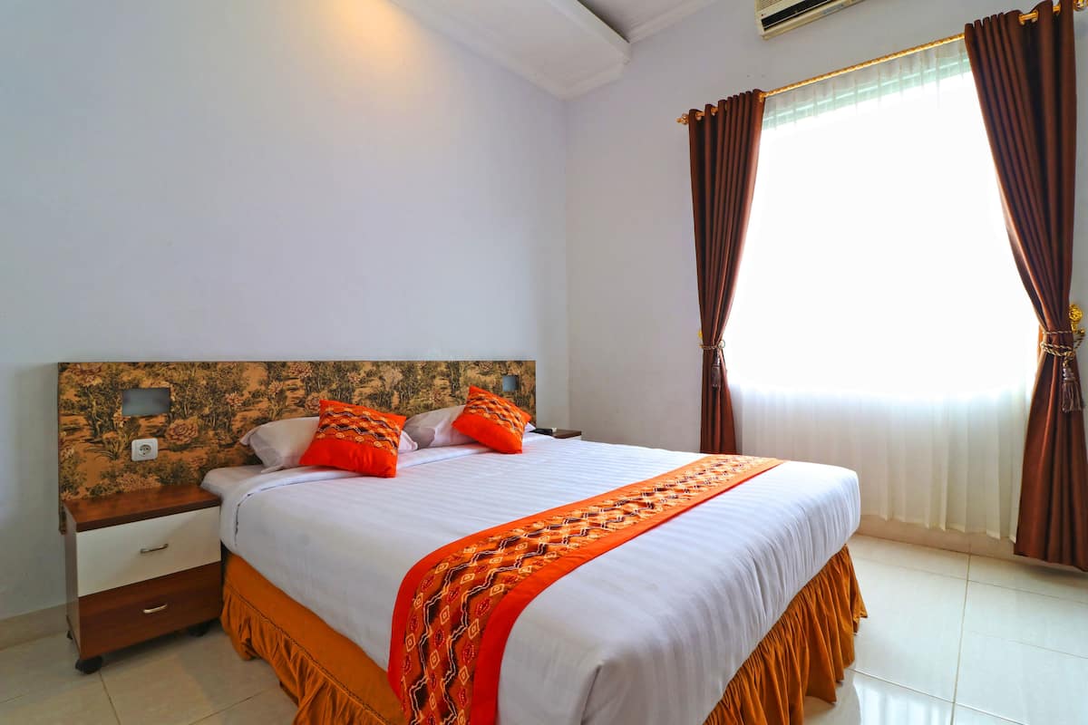 A neatly made bed with a patterned bedspread and pillows, a wooden nightstand with a drawer, and a window with curtains.