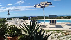 Zona de piscina amb gandules i ombrelles, envoltada de plantes en testos i una vista despejada del mar.