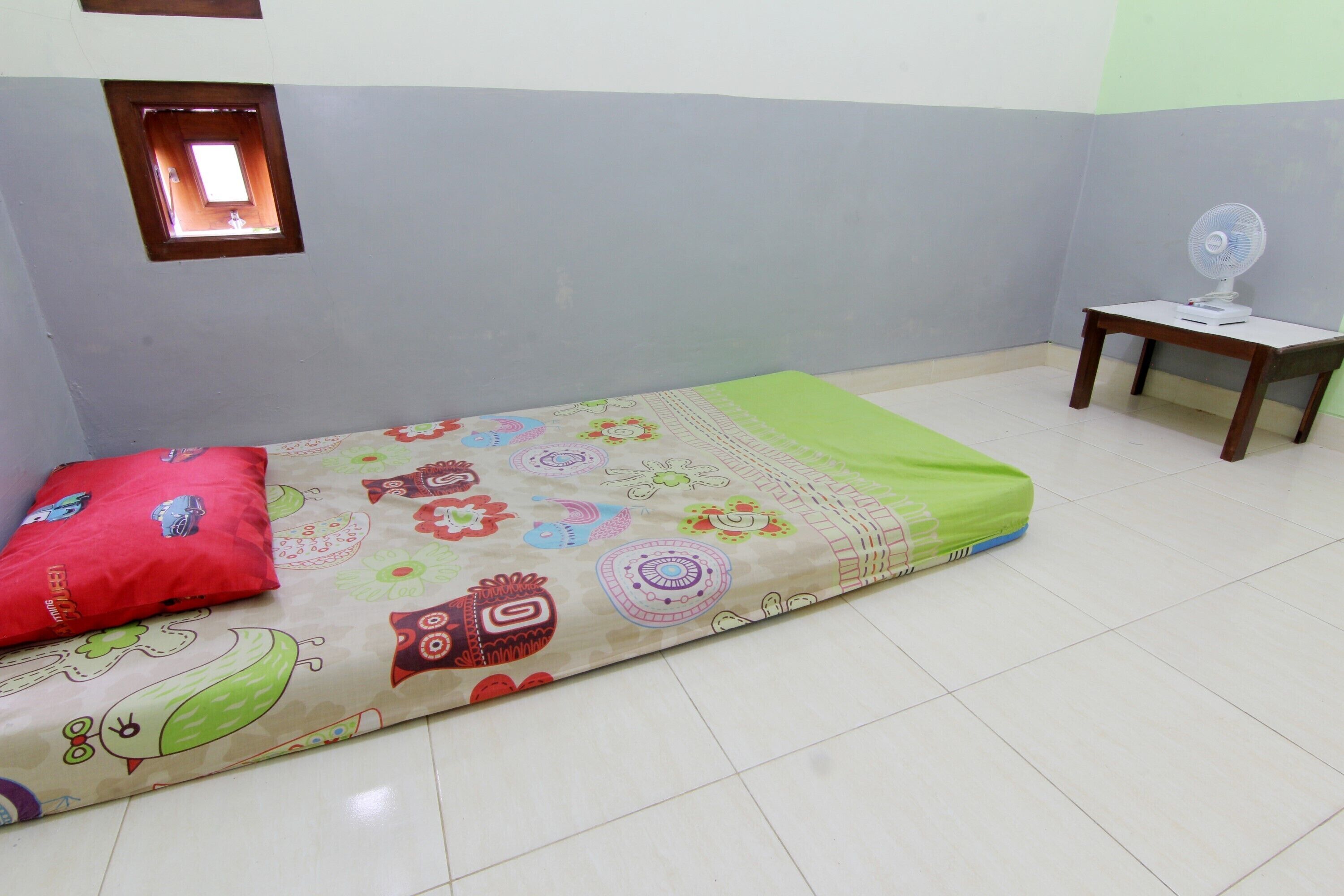 A room with a bed, a small table with a fan, and a window with a view.
