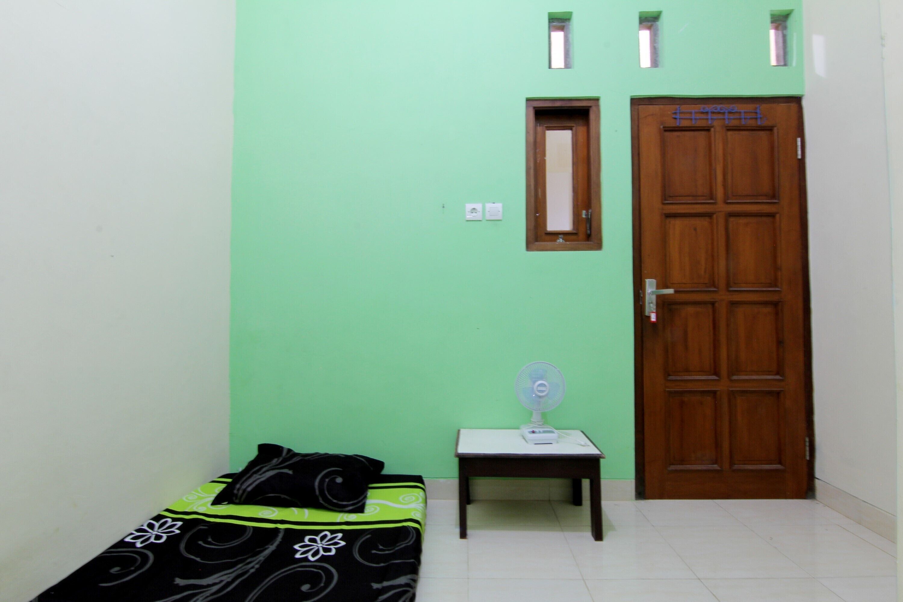 A small room with a bed, a wooden door, a small table with a fan, and a window.
