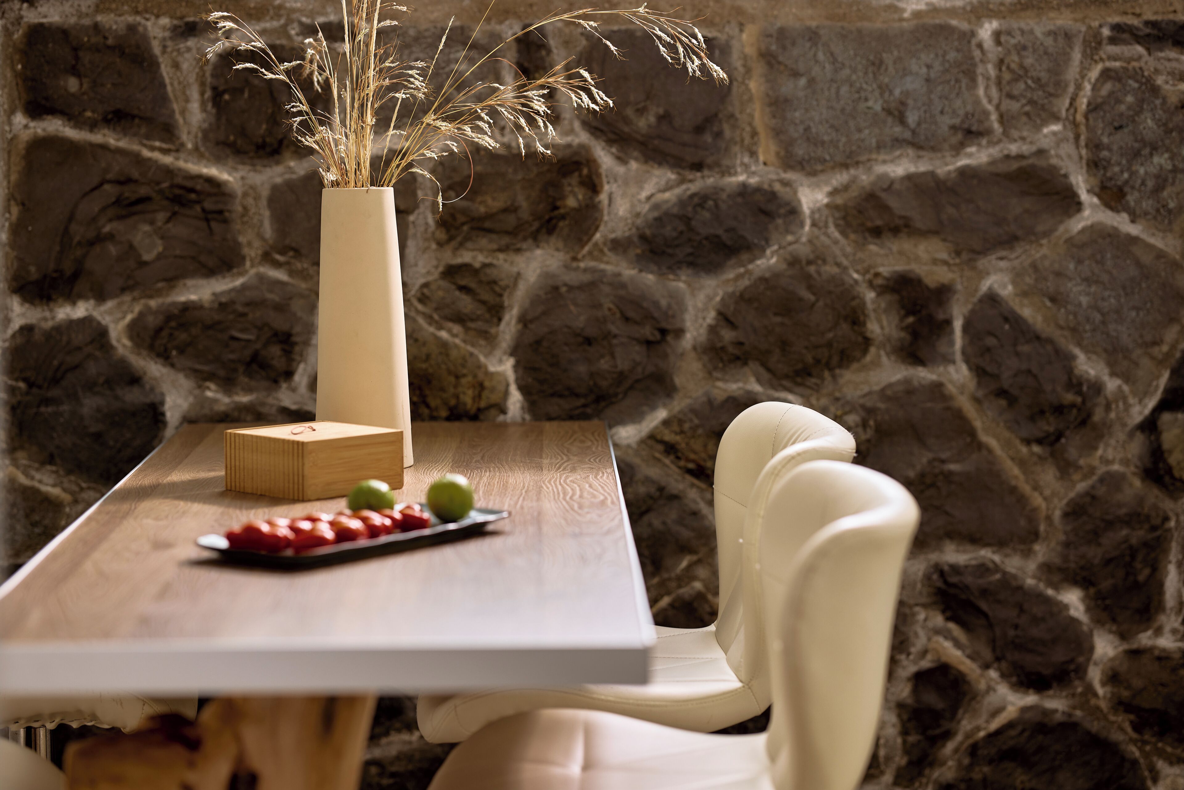 A stone wall, a wooden table with a vase, a wooden box, and a plate with fruit.