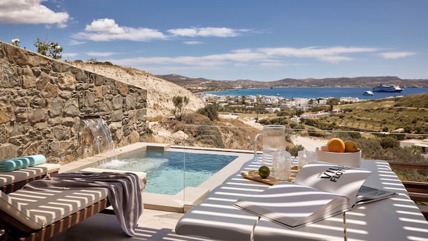 Une piscine bordée d’un muret en pierre, des chaises longues, une table avec un livre et des fruits, et une vue sur la mer et les collines.