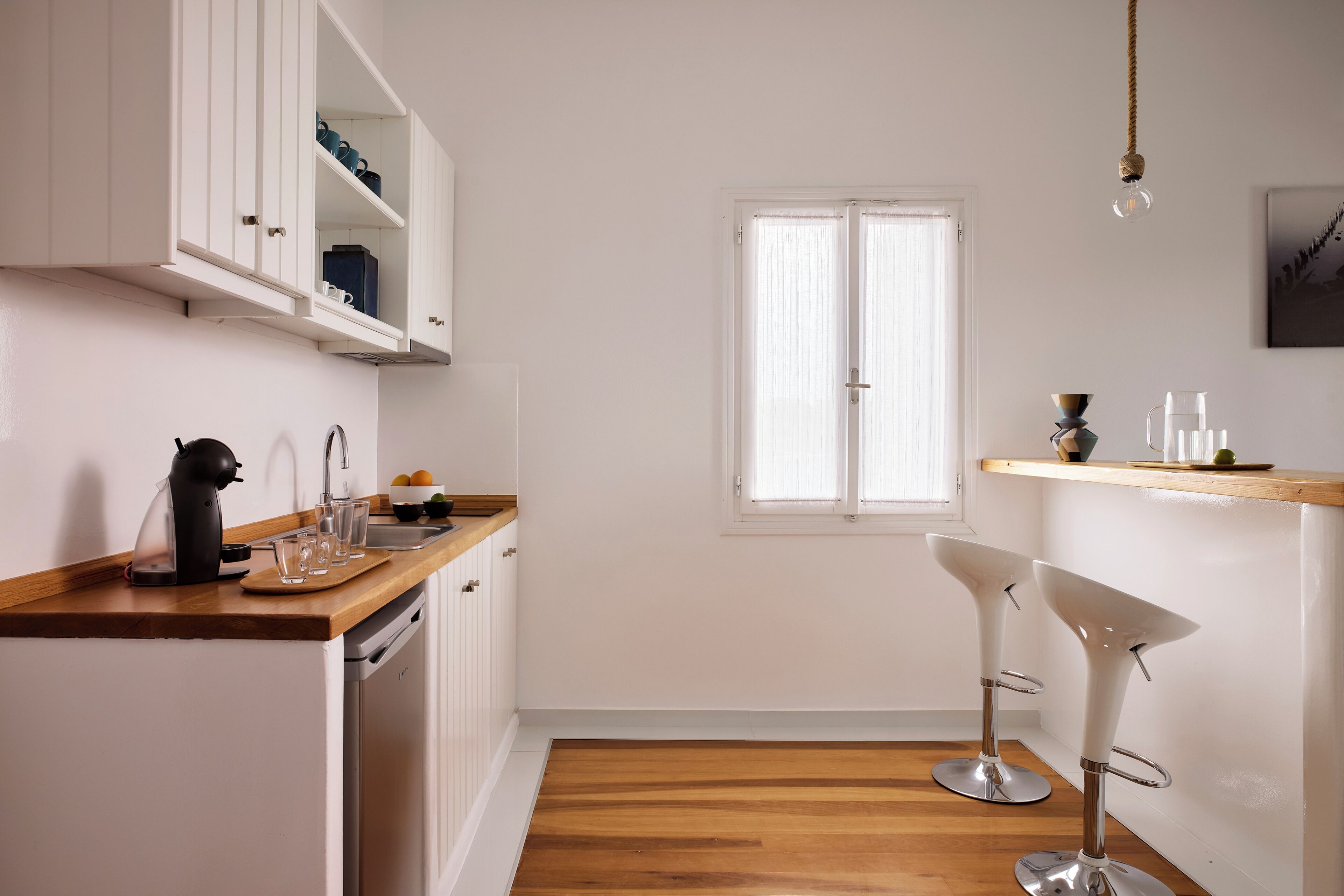 A kitchen with white cabinets, a wooden countertop, a kettle, glasses, and a window.