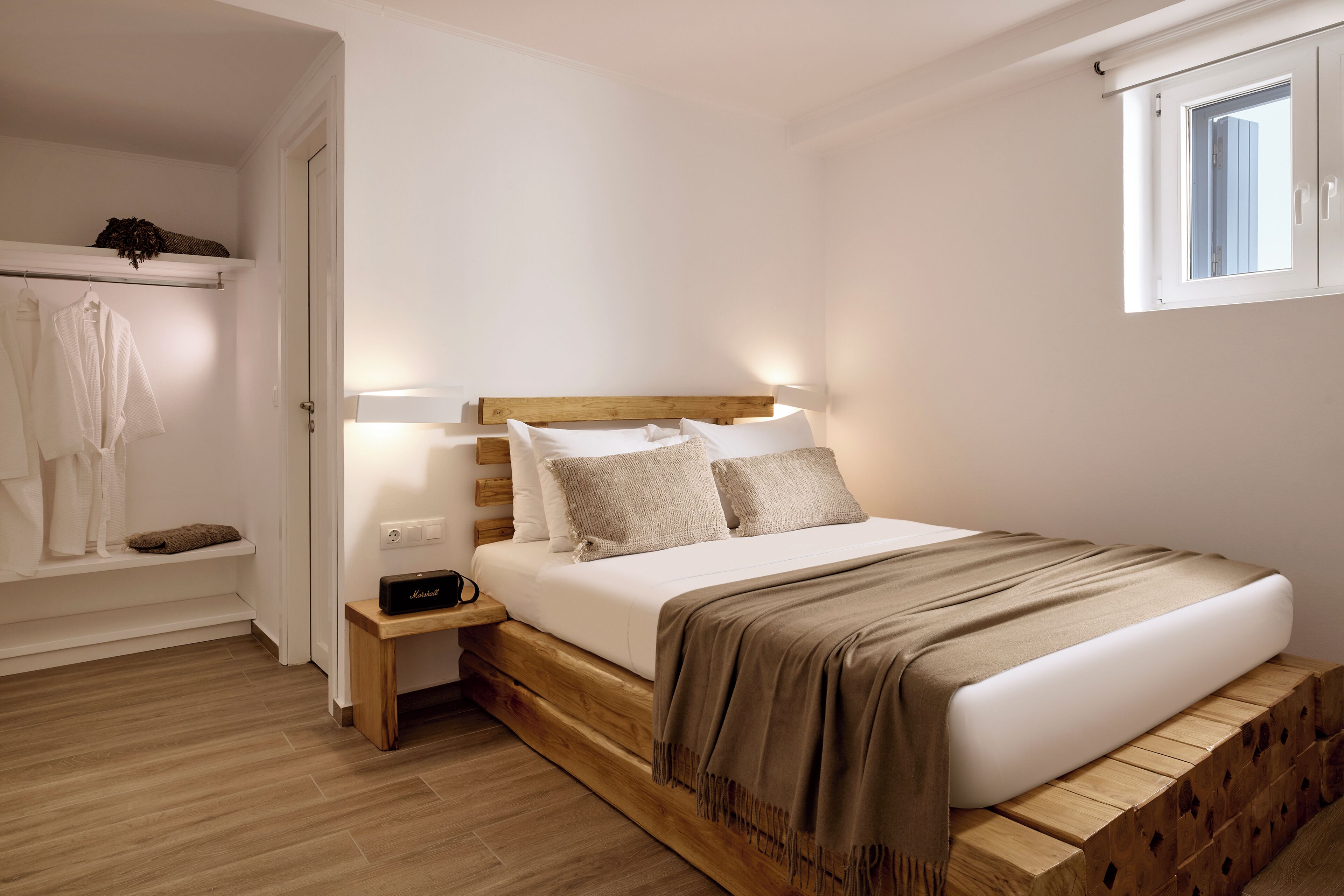 A bedroom with a wooden bed, white walls, a small bedside table, a nightstand, a window, and a built-in shelf with clothes.