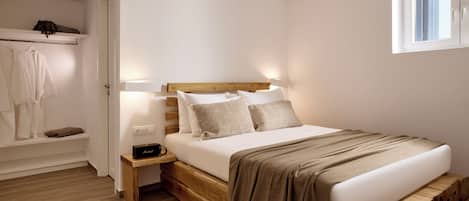 A bedroom with a wooden bed, white walls, a small bedside table, a nightstand, a window, and a built-in shelf with clothes.