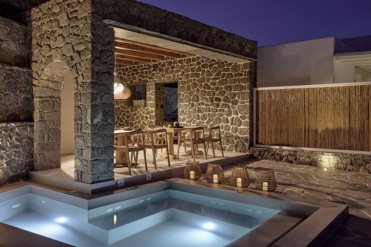 A stone exterior wall, a pool with lights, and a wooden deck with chairs.