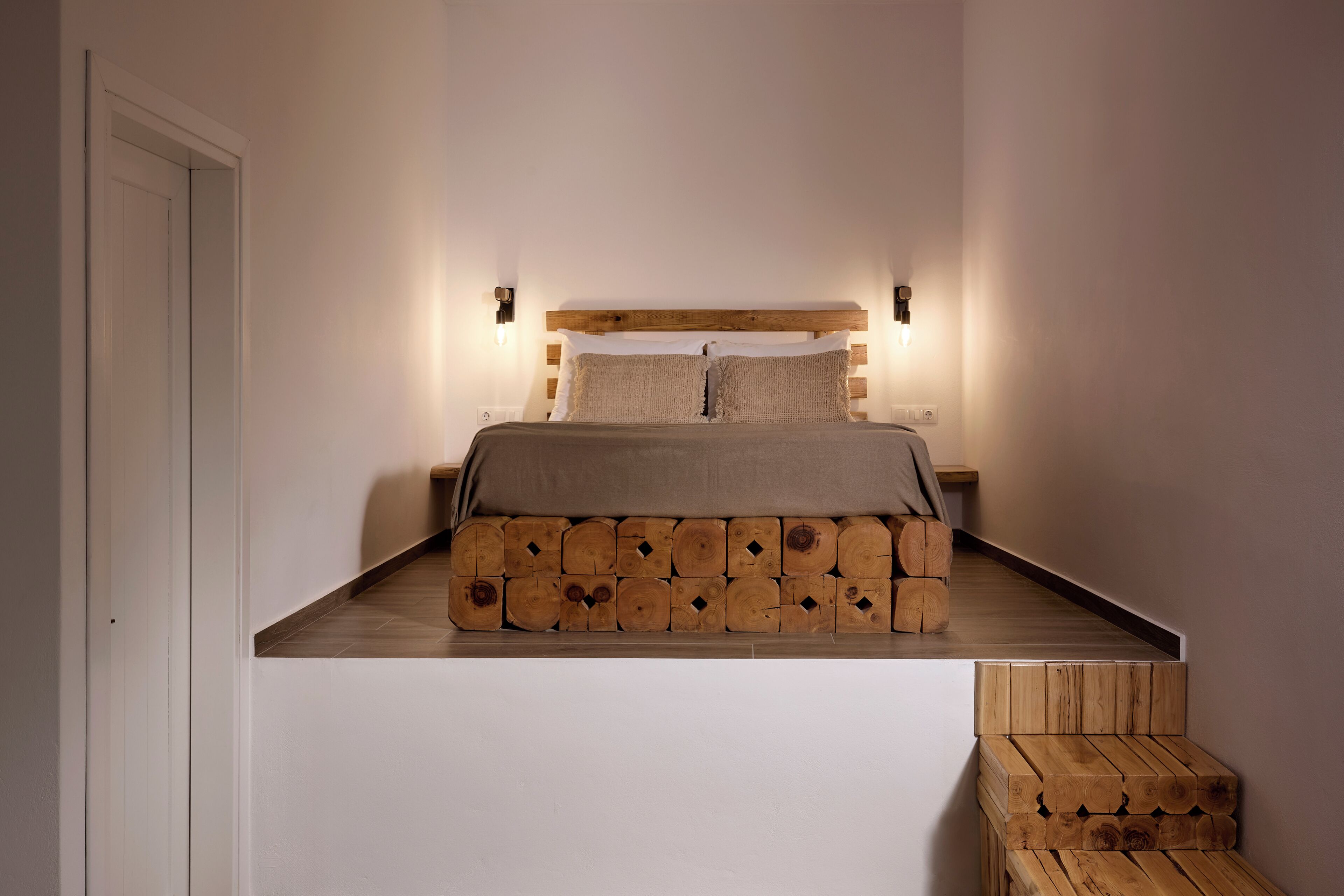 Une chambre à coucher avec un lit sur une plateforme surélevée, une tête de lit en bois et des luminaires fixés au mur.