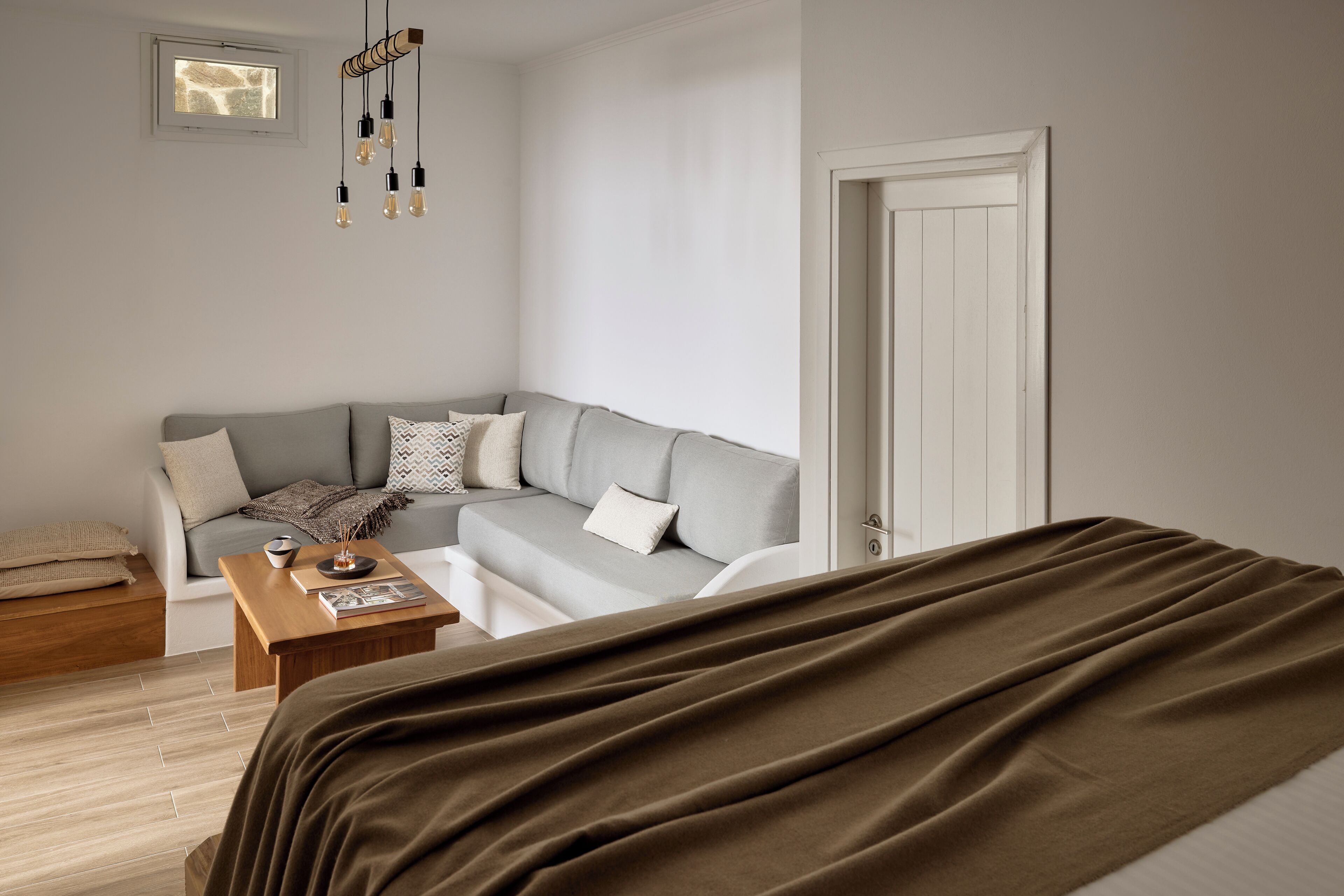 A modern bedroom with a grey sofa, a wooden coffee table, a bed with a brown blanket, and a white door.