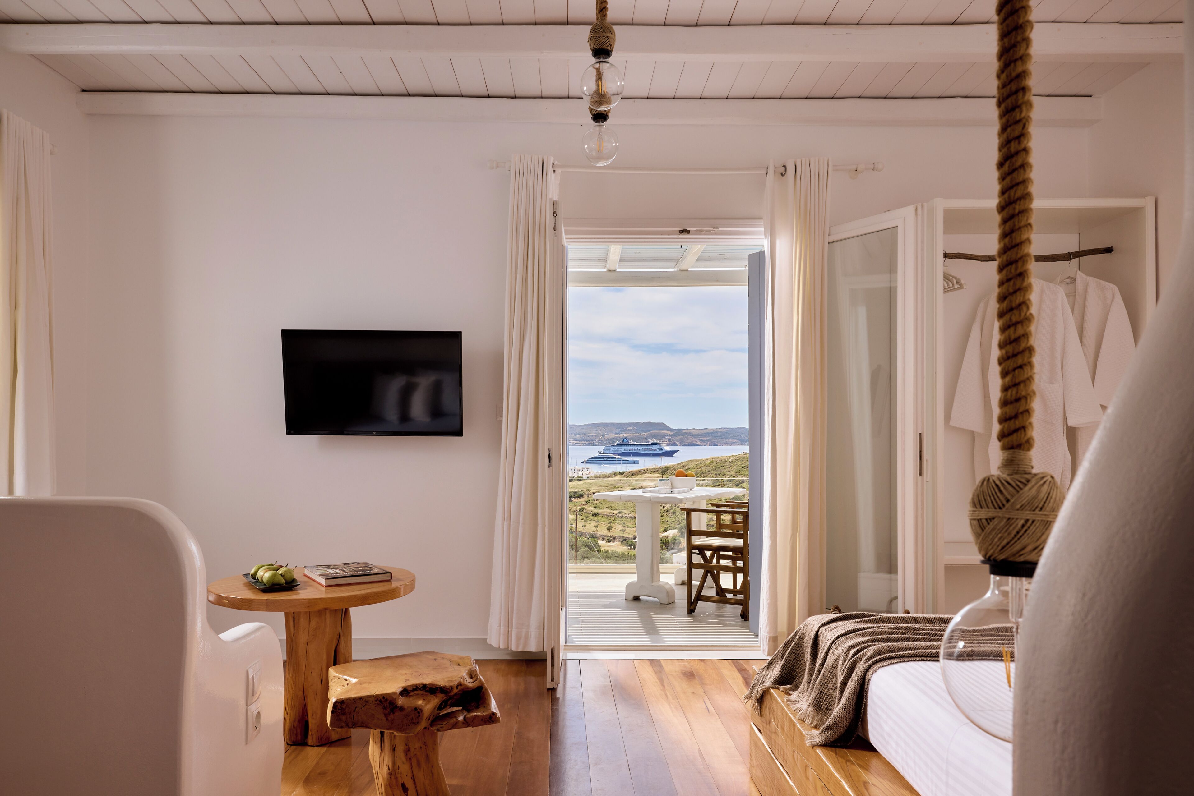 A room with a bed, a TV mounted on the wall, a wooden table, and a view of the sea through an open door.