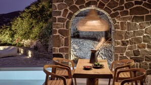 Un mur en pierre avec une cheminée, une piscine et des chaises en bois disposées autour d’une table.
