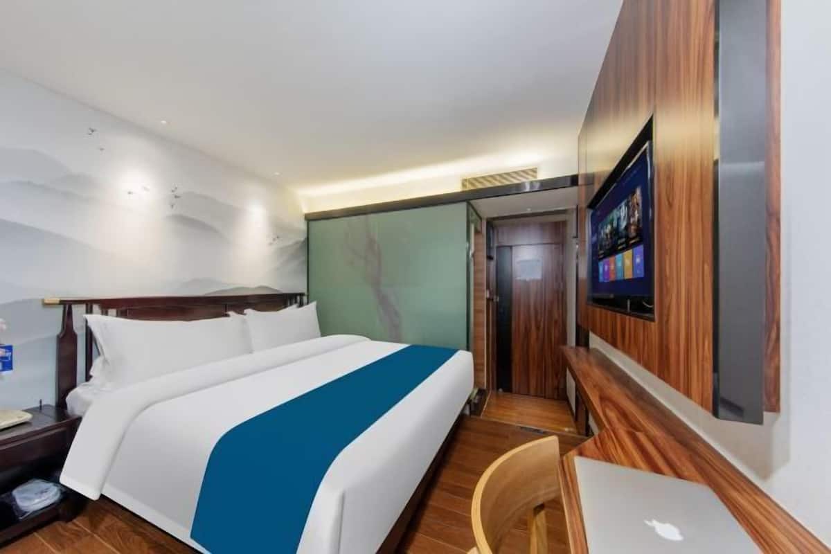 A hotel room with a bed, a desk with a laptop, a TV mounted on the wall, and a door leading to another room.