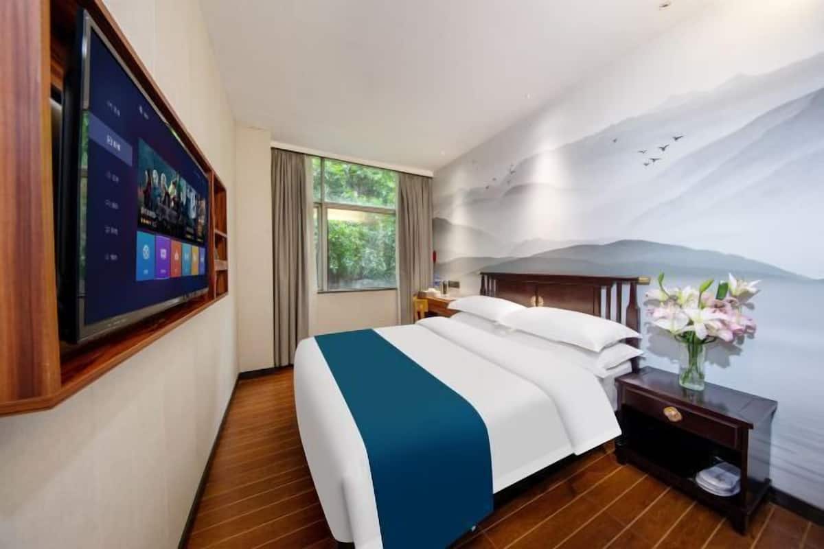 A hotel room with a bed, a TV mounted on the wall, a nightstand, and a window with curtains.