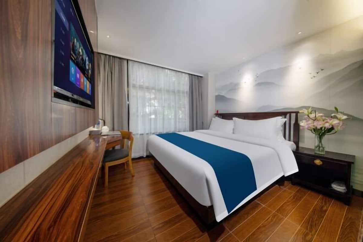 A hotel room with a large bed, a desk, a chair, a TV mounted on the wall, and a nightstand with a vase of flowers.