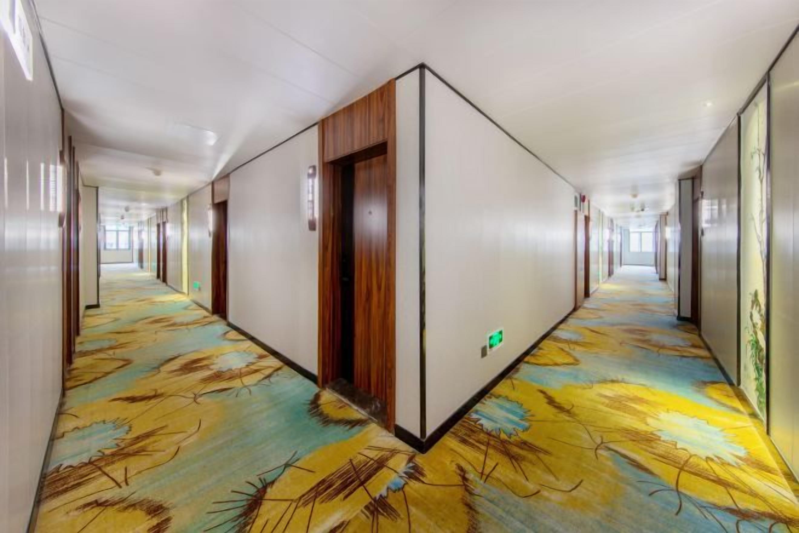 A hotel corridor with a patterned carpet, wooden doors, and a green exit sign.